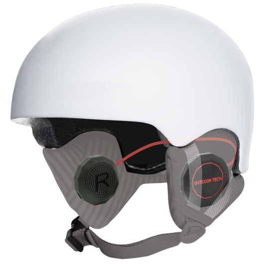 A white ski or snowboard helmet with integrated dark visor lenses and gray ear cushions. One ear cushion has a visible circular badge reading 'OUTDOOR TECH' with a red outline, and the other shows a dark green ear plug with a white 'R'.