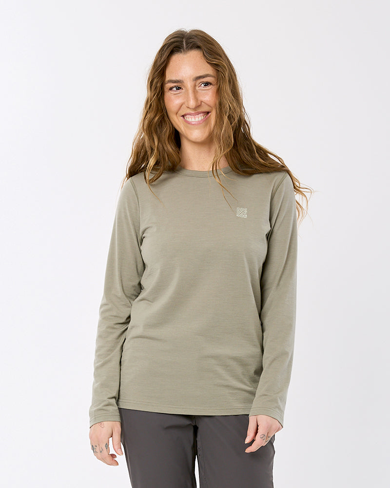 A woman with long light brown hair smiling, wearing a long-sleeve crewneck shirt in a muted sage green color, paired with dark gray pants, against a plain white studio background. XTM merino wool base layer thermal top women womens snow cold khaki