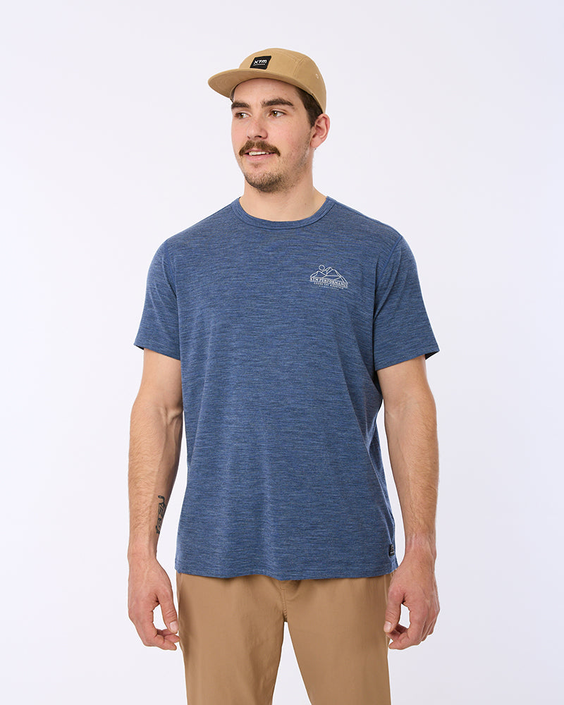 A man wearing a heathered blue crewneck t-shirt with a small white chest graphic, paired with tan pants and a tan baseball cap, standing against a light gray studio background. XTM wool merino indigo t-shirt base layer thermal mens