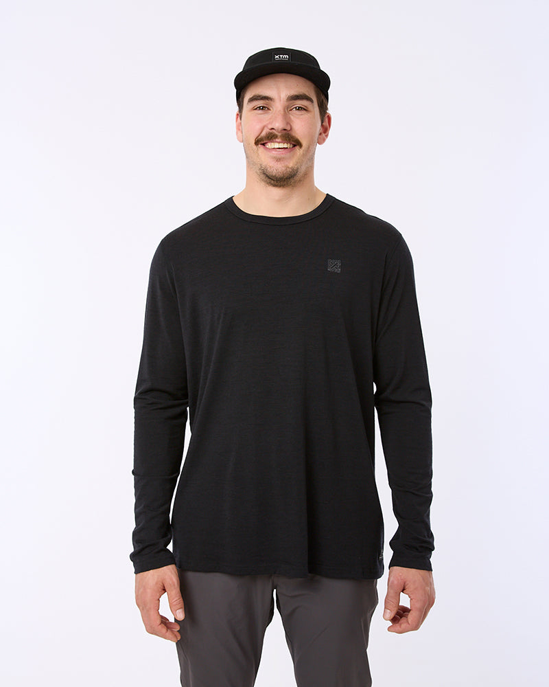 A man wearing a black long-sleeve crewneck shirt, dark gray pants, and a black baseball cap, standing against a plain white studio background. XTM black icon merino wool tencel thermal top mens