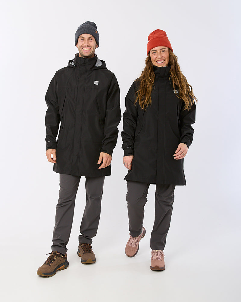 A man and a woman modeling matching black hooded jackets and dark gray pants against a plain light gray studio background. The man wears a dark beanie and brown shoes, while the woman wears a red beanie and pinkish-brown shoes. XTM Innisfail  mens womens rain coat waterproof 